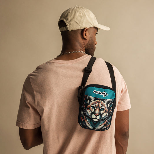Front view of Manly Cougar Utility Crossbody Bag thrown over left shoulder of a male, bag in turquoise blue and black with head shot of a cougar and text "manly" at top