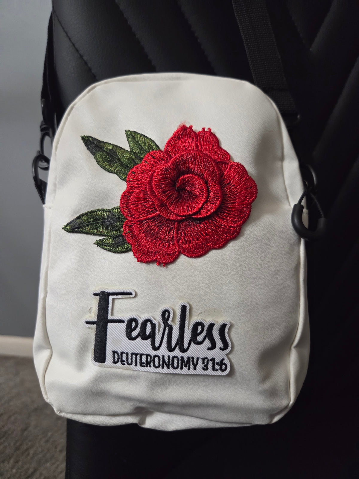 A white cross body bag with a red rose patch and a fearless patch, shown resting on a black chair with a blue strap visible. Close up view.