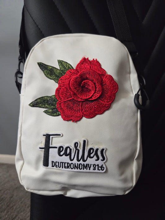 A white cross body bag with a red rose patch and a fearless patch, shown resting on a black chair with a blue strap visible. Close up view.