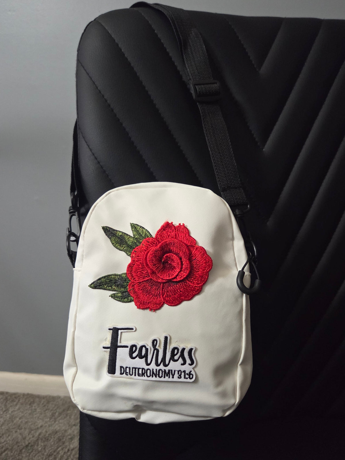 A white cross body bag with a red rose patch and a fearless patch, shown resting on a black chair with a blue strap visible. Distant view.