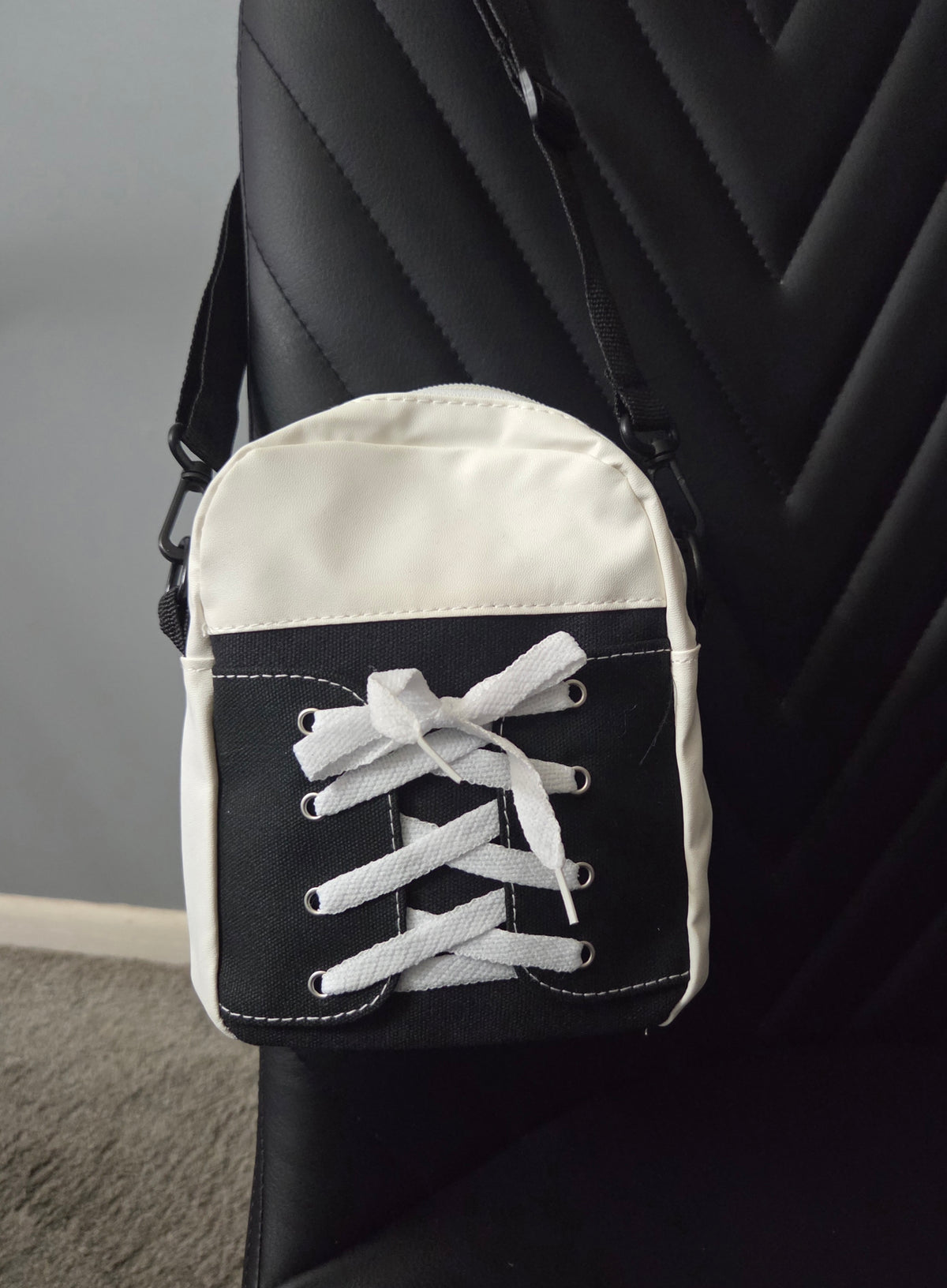 Back view of Black and White Sneaker cross body bag in faux leather and cloth blend fabrics, featuring a black strap, resting on a black leather chair. Close up view.