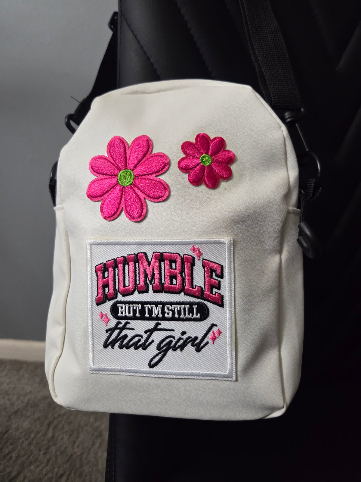 A white cross body bag with colorful embroidered flowers and "humble" and patches, shown resting on a black chair with a black strap visible. Close up view.