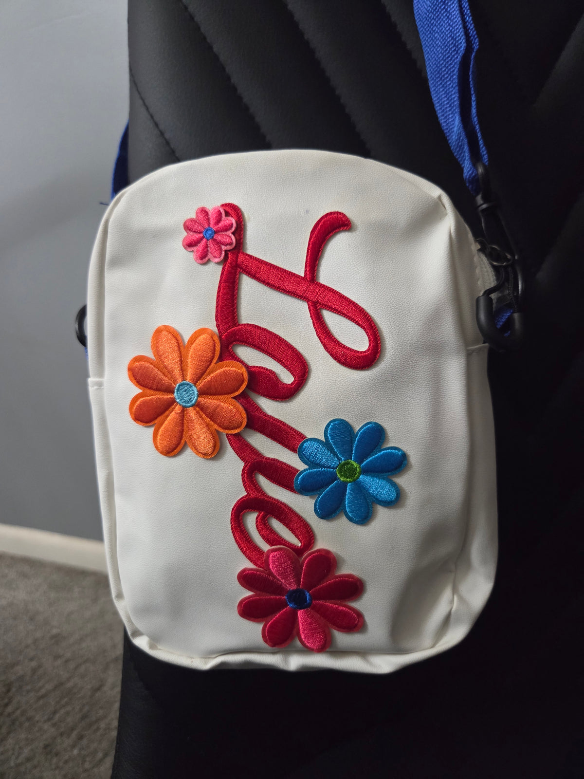A white cross body bag with colorful embroidered floral and script designs, shown resting on a black chair with a blue strap visible. Close up view.