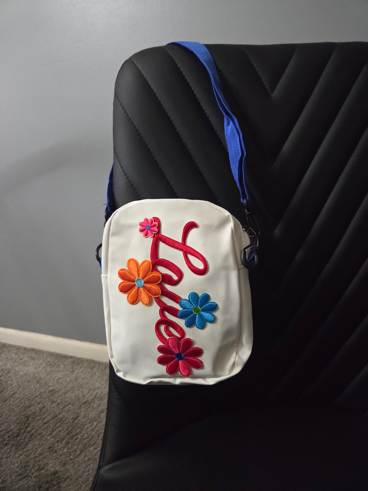 A white cross body bag with colorful embroidered flowers and a red script design, featuring a blue strap, resting on a black leather chair. Distant view.