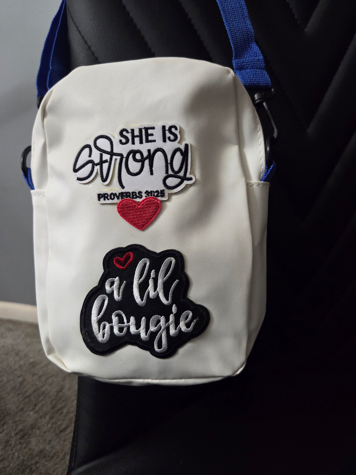 A white crossbody bag with patches that I am strong diva, shown resting on a black chair with a blue strap visible. Close up view.