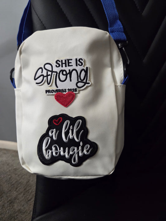 A white crossbody bag with patches that I am strong diva, shown resting on a black chair with a blue strap visible. Close up view.