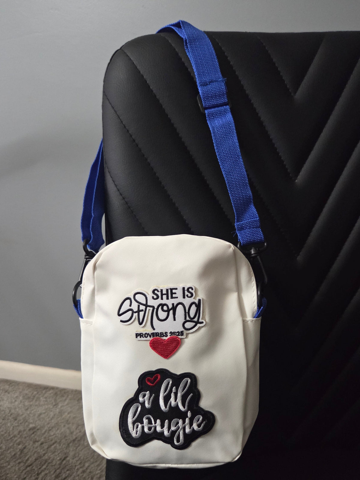 A white crossbody bag with patches that say I am strong diva, shown resting on a black chair with a blue strap visible. Distant view.
