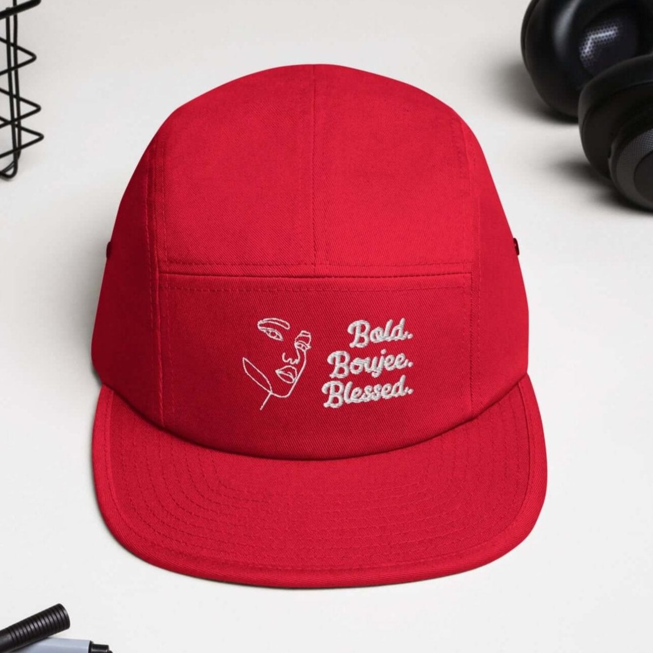 Front view of a red five-panel cap on a white desk with white text saying 'Bold, Boujee, Blessed'