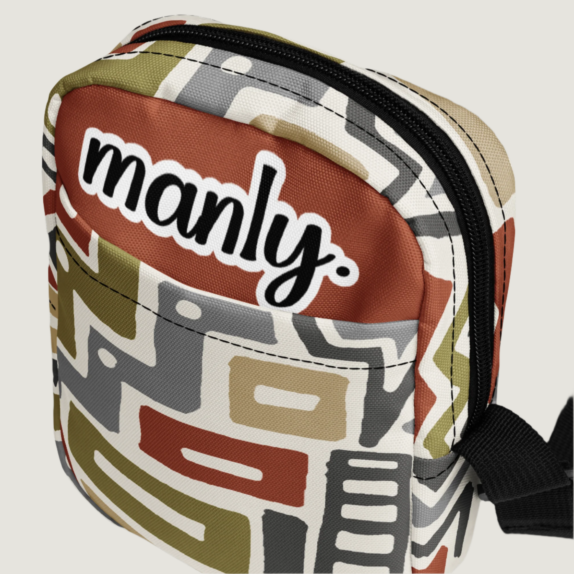 Front slanted view of Manly Tribal Tune Utility Crossbody Bag in beige and tan print text "manly" at top