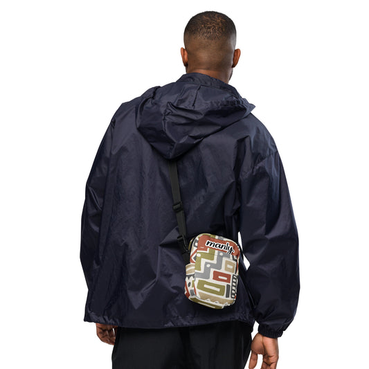 Back view of Manly Tribal Tune Utility Crossbody Bag being worn by a male, bag in beige and tan print text "manly" at top