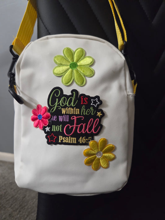 A white crossbody bag with colorful embroidered flowers and Psalm 46:5 scripture...God is with her...script, shown resting on a black chair with a yellow strap visible. Close up view.