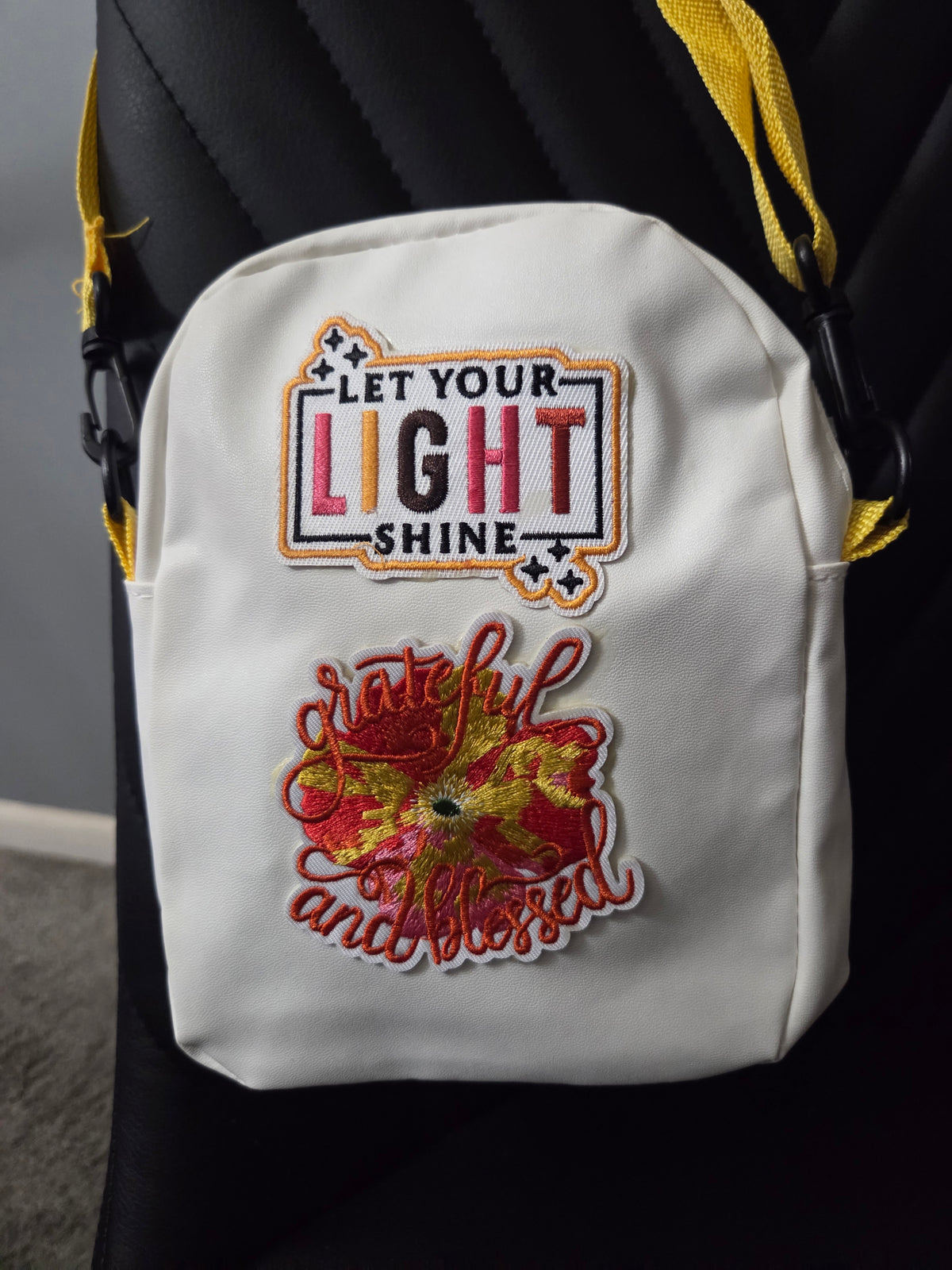 A white cross body bag with colorful embroidered "let your light shine" and "grateful and blessed patches, shown resting on a black chair with a yellow strap visible. Close up view.