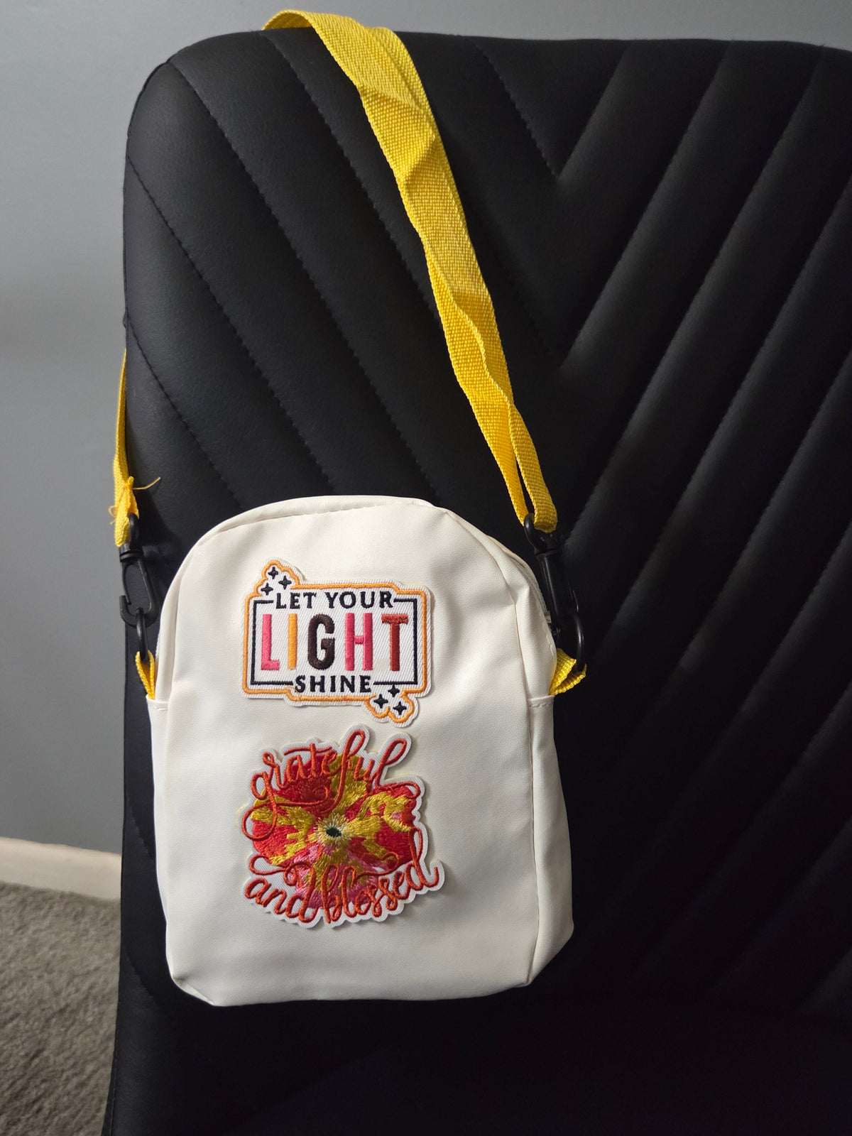 A white cross body bag with colorful embroidered "let your light shine" and "grateful and blessed patches, shown resting on a black chair with a yellow strap visible. Distant view.